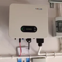 Zamontowany biały inwerter SOFAR, obok skrzynka bezpiecznikowa i licznik energii, połączone białymi rurami PCV na białej ścianie, widoczny fragment okna.