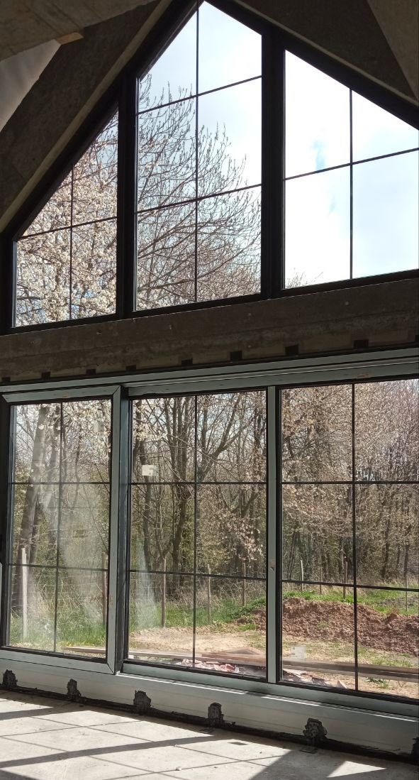 Duże okno z czarnymi szprosami w surowym wnętrzu, widok na kwitnące drzewa na zewnątrz.