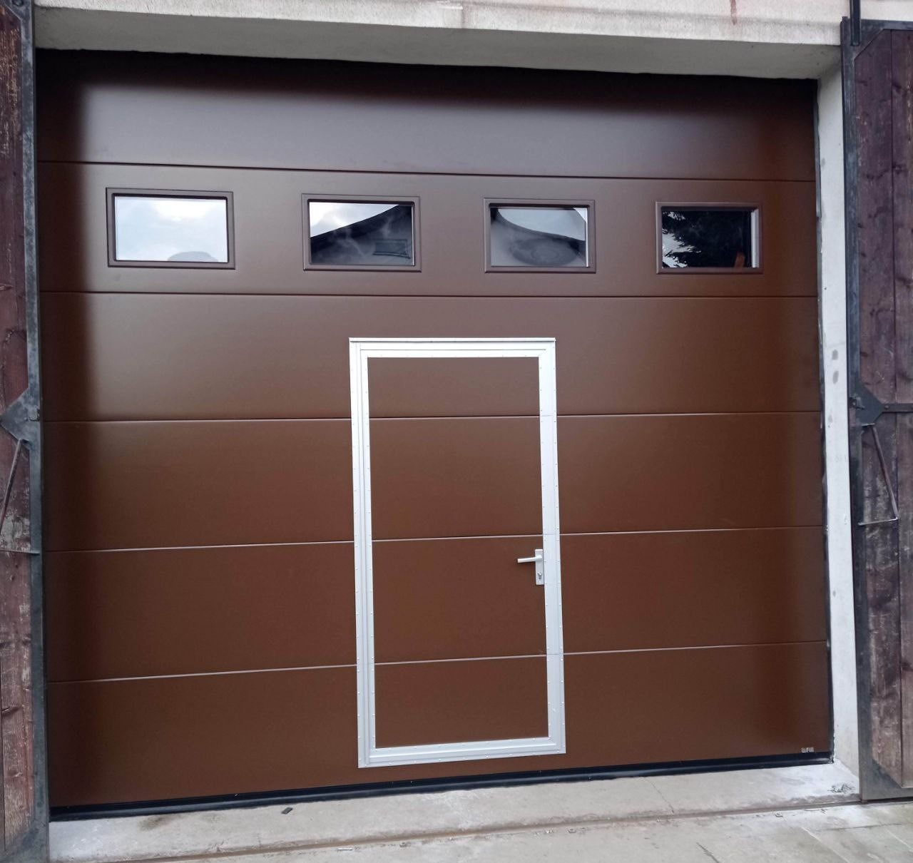Brązowa segmentowa brama garażowa z czterema małymi okienkami i drzwiami wejściowymi pośrodku, zamontowana w betonowym garażu.