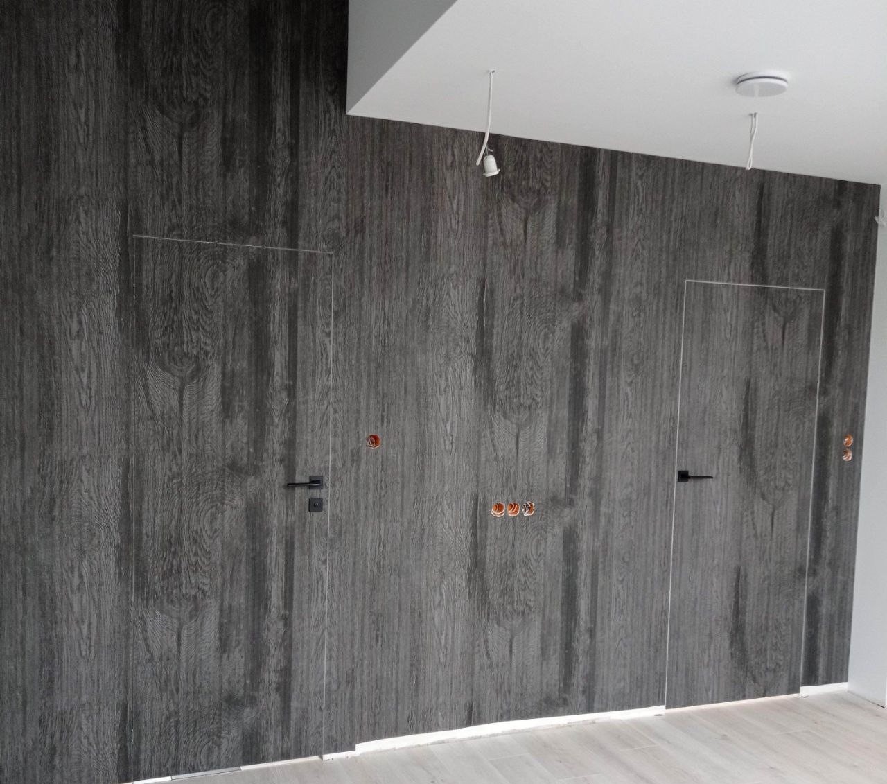 Ściana z szarego panela imitującego drewno z dwoma ukrytymi drzwiami z czarnymi klamkami i wystającymi przewodami elektrycznymi.