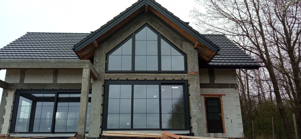 Surowy stan domu jednorodzinnego w trakcie budowy z zamontowanymi oknami w czarnych ramach o dużej powierzchni przeszkleń i nieregularnym kształcie, widoczne bloczki betonowe i elementy konstrukcyjne.