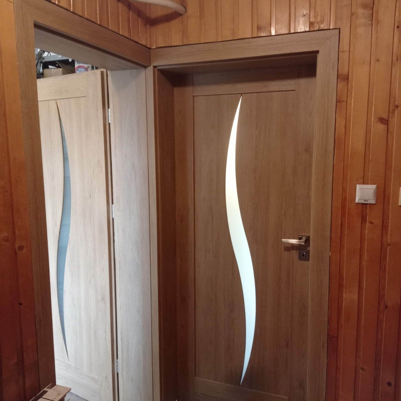 Dwa drewniane skrzydła drzwiowe z dekoracyjnymi, falistymi przeszkleniami, zamontowane w drewnianej ościeżnicy, widoczne na tle boazerii.