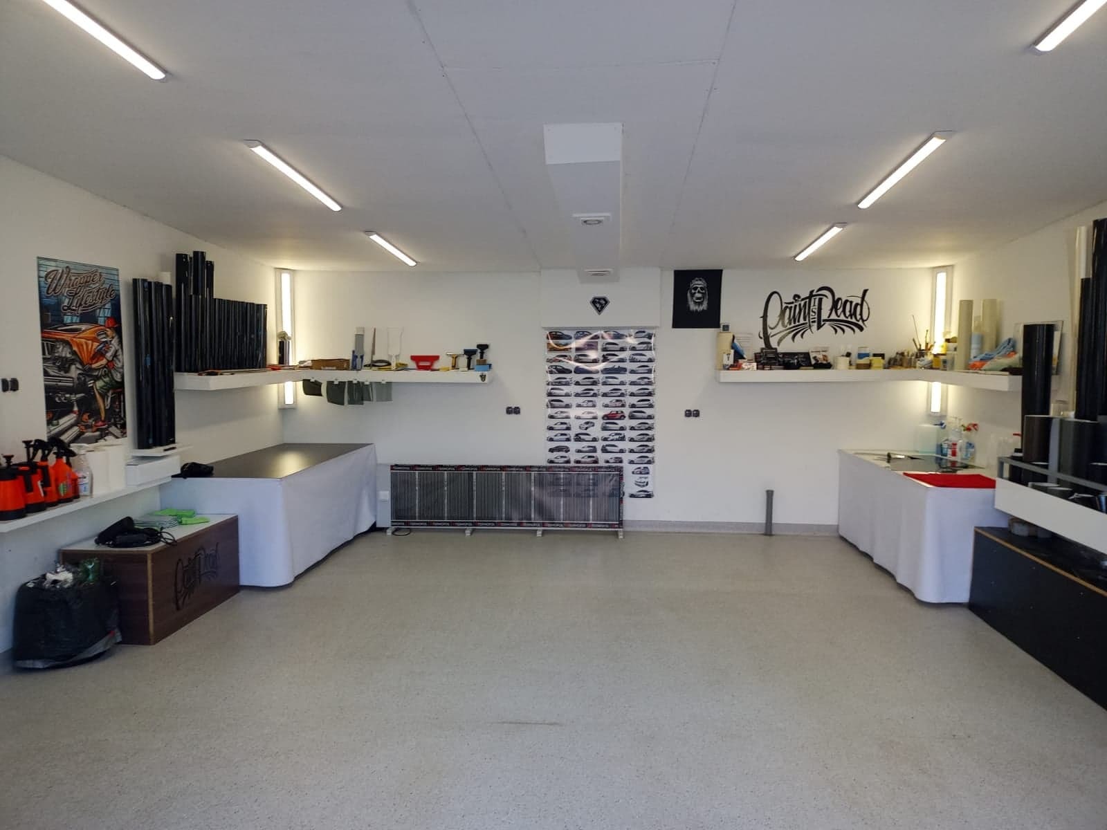 Przestronne, dobrze oświetlone studio z przygotowanym stanowiskiem pracy, rolkami folii i narzędziami na półkach, plakatami i listą modeli samochodów na ścianach, gotowe do pracy nad pojazdami.