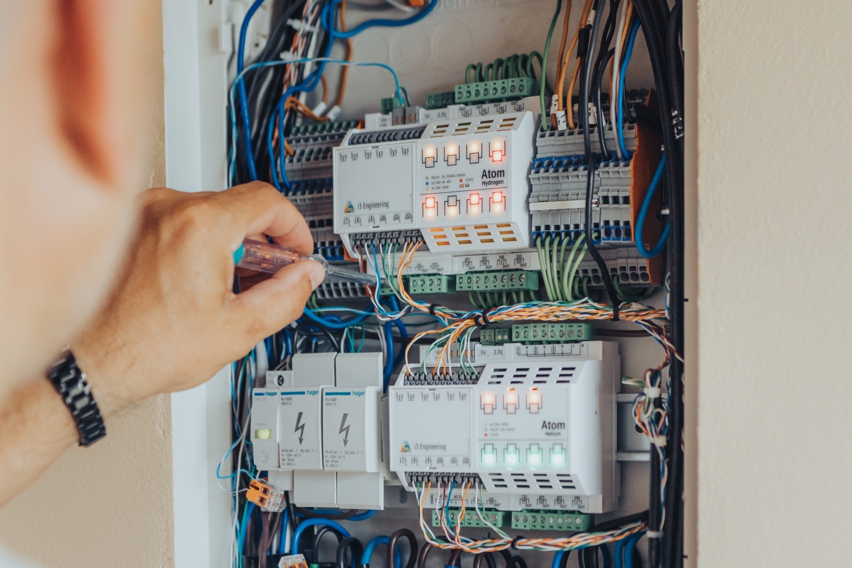 Elektryk z testerem napięcia sprawdza połączenia w rozdzielnicy z modułami Atom Hydrogen i Atom Helium, widoczne przewody, złączki i zabezpieczenia Hager.