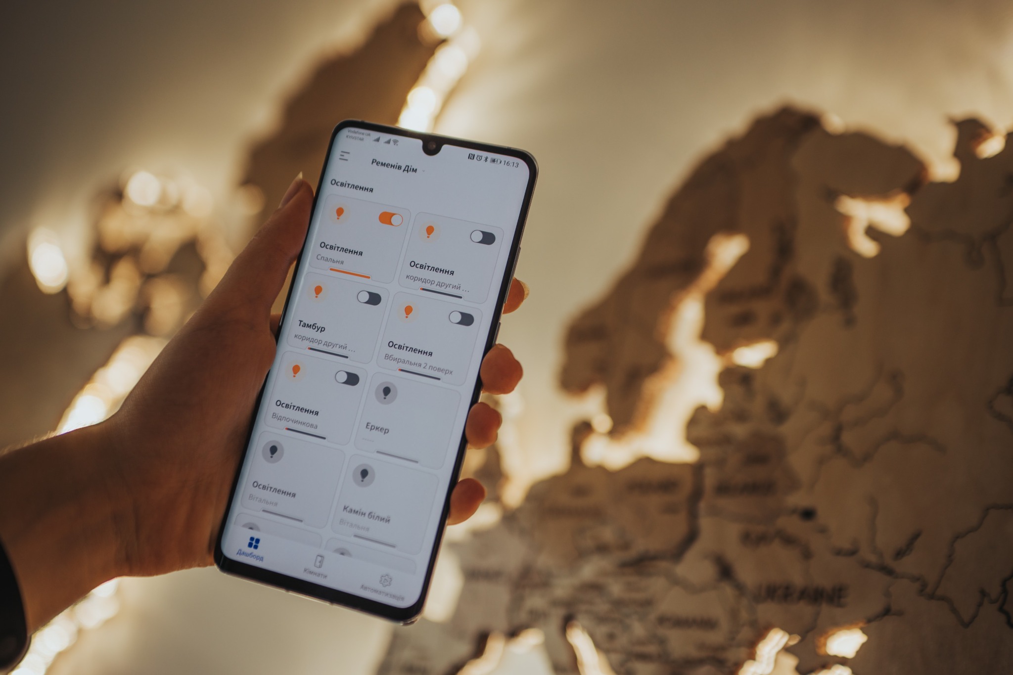 Smartfon w dłoni wyświetlający aplikację do sterowania oświetleniem w domu, z mapą Europy w tle.