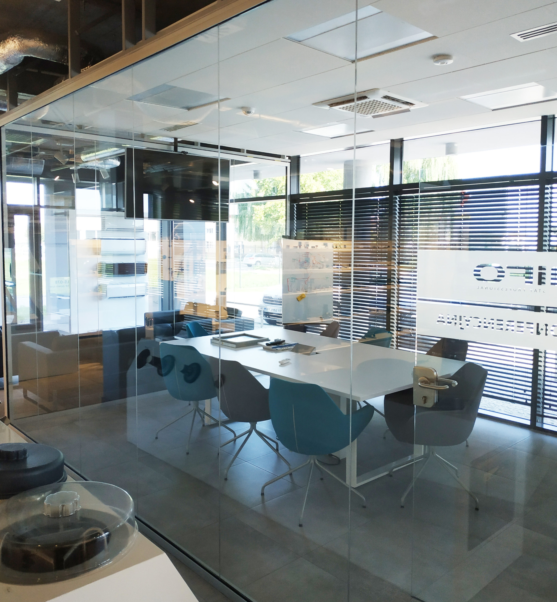 Szklana sala konferencyjna z białym stołem, krzesłami w odcieniach szarości i błękitu, widok przez szybę na zewnątrz, na ścianie logo firmy za żaluzjami.