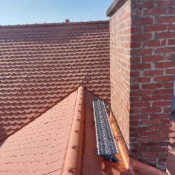 Ceglasty dach z ceramicznej dachówki karpiówki, z zamontowaną metalową ławą kominiarską przy ceglanym kominie na tle błękitnego nieba.