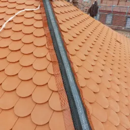 Montaż taśmy kalenicowej Braas Figaroll Plus na dachu z dachówki karpiówki, widoczny pracownik w kombinezonie roboczym i rusztowanie w tle.