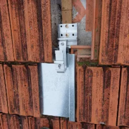 Szczegółowy widok mocowania szyny K2 do dachówki ceramicznej, z widocznym fragmentem łaty i kontrłaty oraz butem roboczym w dolnej części kadru.