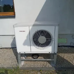 Biała pompa ciepła Stiebel Eltron zamontowana na metalowej konstrukcji na zewnątrz budynku z oknem, widoczne przewody i kamieniste podłoże.