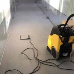 Profesjonalny odkurzacz piorący koloru żółto-czarnego stoi na szarym dywanie w długim korytarzu biurowym z przeszklonymi ścianami. Widoczny kabel zasilający podłączony do gniazda w podłodze.