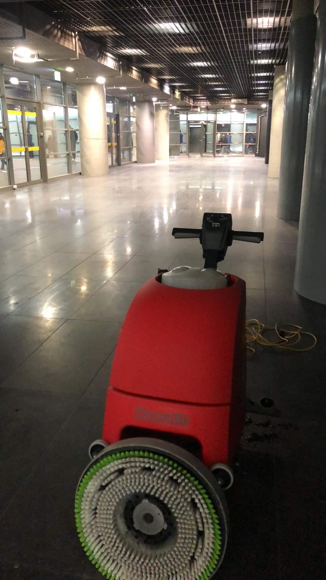 Czerwona maszyna czyszcząca z obrotową szczotką na posadzce w przestronnym holu z kolumnami i szklanymi drzwiami wejściowymi w tle.
