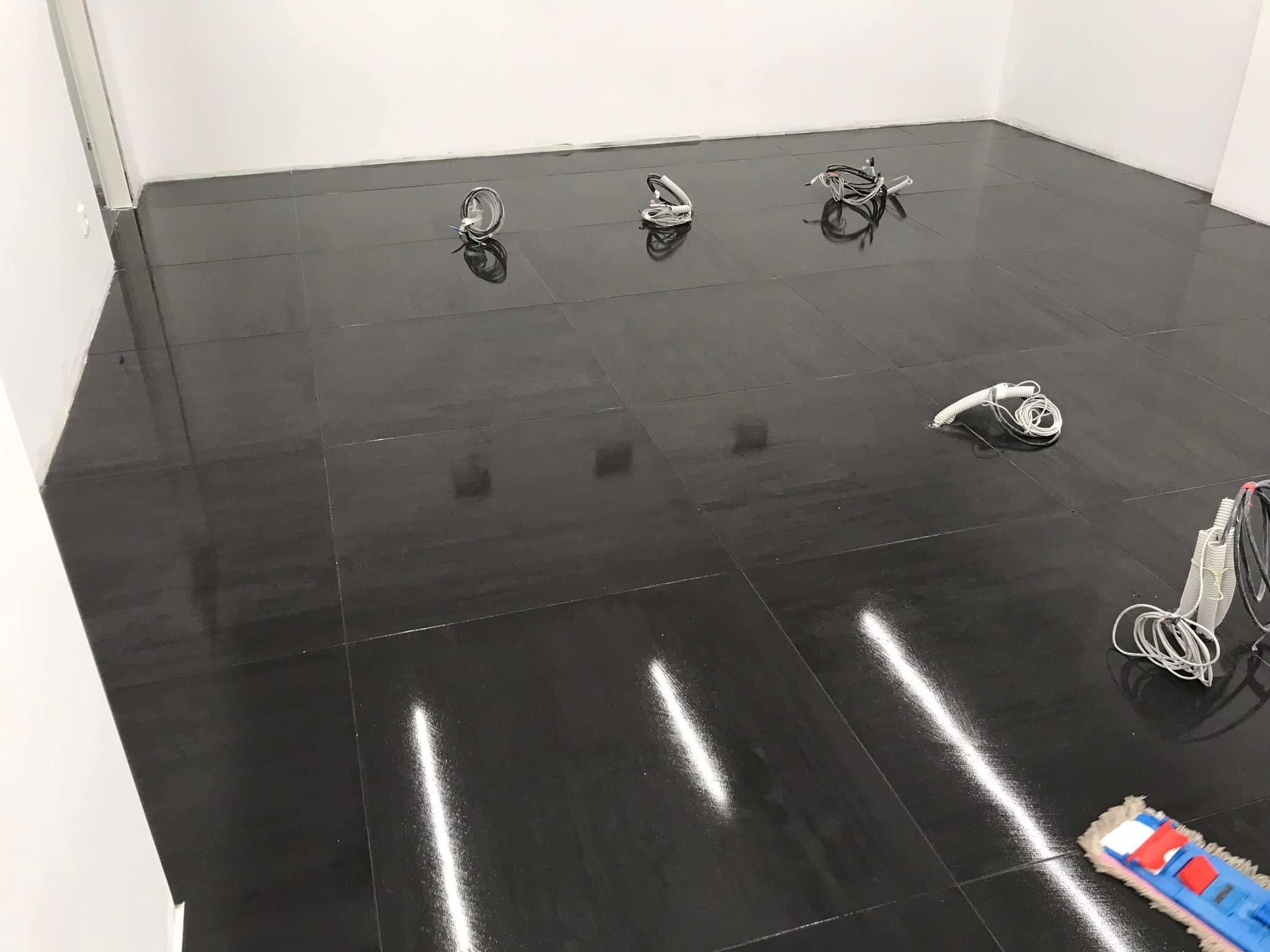 Lśniąca, świeżo umyta podłoga z ciemnych płytek w pustym pomieszczeniu biurowym, widoczne kable i mop na podłodze, ślady po wodzie.