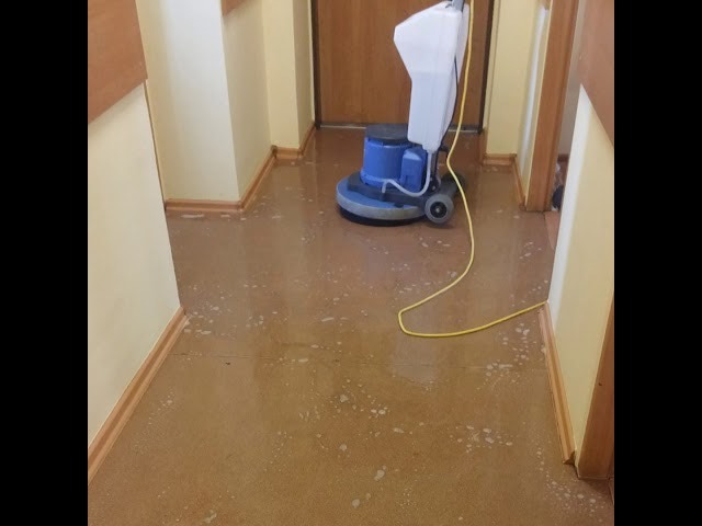 Maszyna do czyszczenia podłóg w trakcie pracy na błyszczącej, brązowej posadzce w korytarzu biurowym, widoczne drobne zabrudzenia i piana.