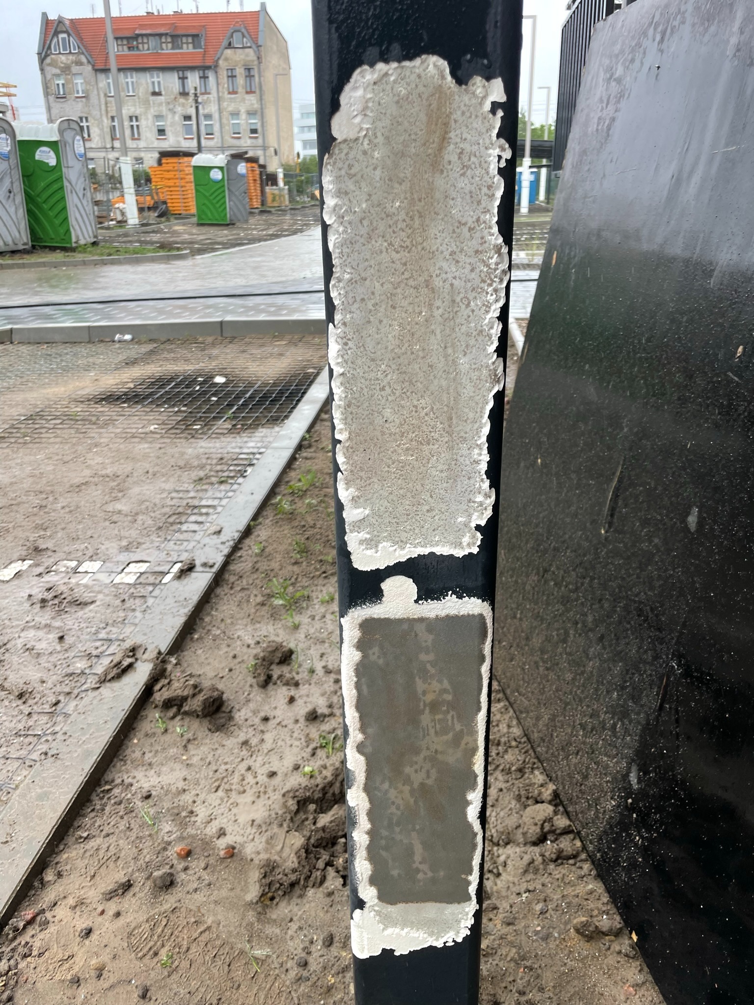 Zniszczona, odchodząca farba z metalowego słupka w Łopusznie, widoczne ślady korozji i resztki starej powłoki, w tle budynek i teren budowy.