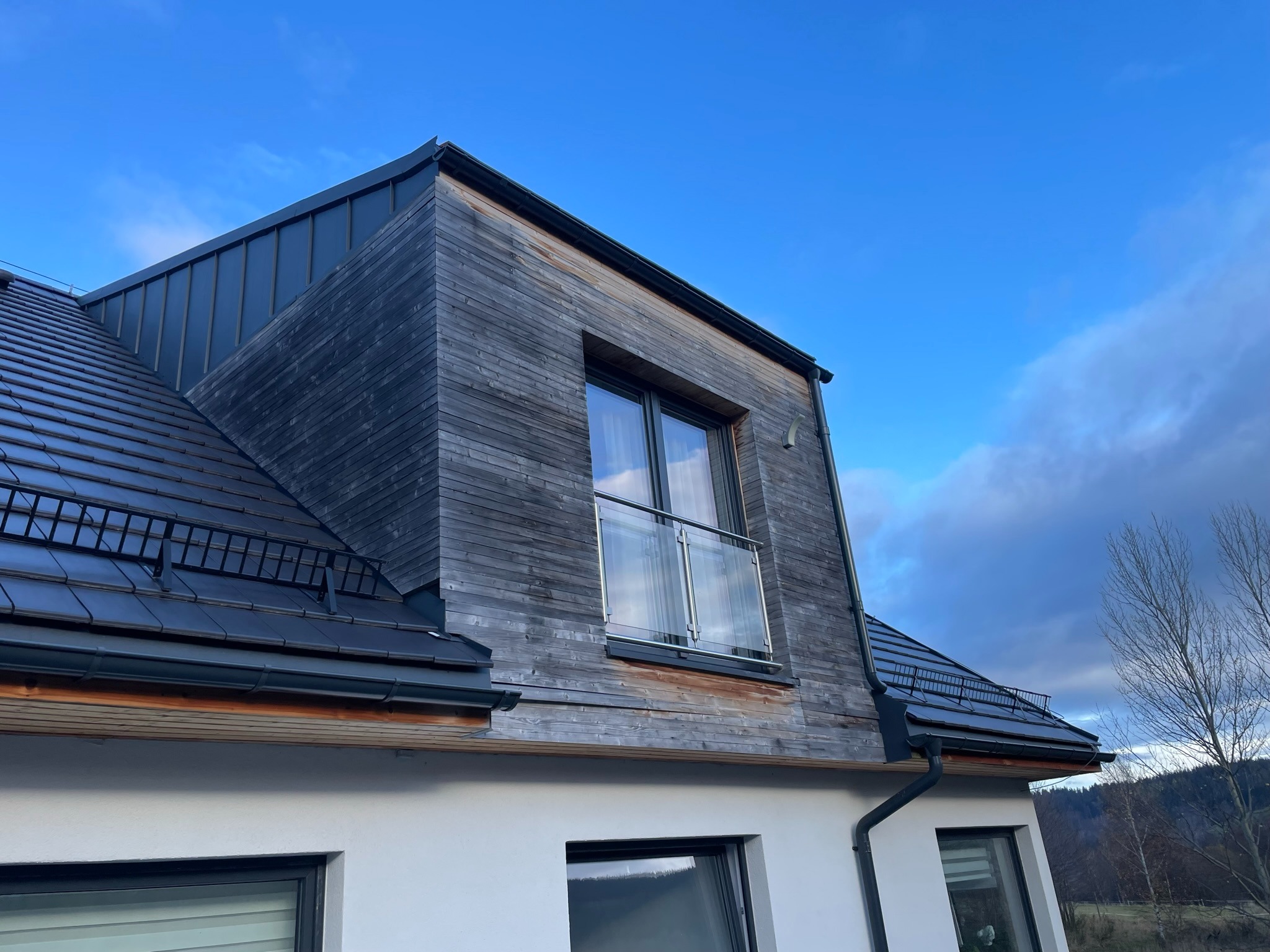 Nowoczesny dom z szarego drewna z balkonem z szklaną balustradą, czarną dachówką i rynnami, widok z dołu na tle błękitnego nieba.