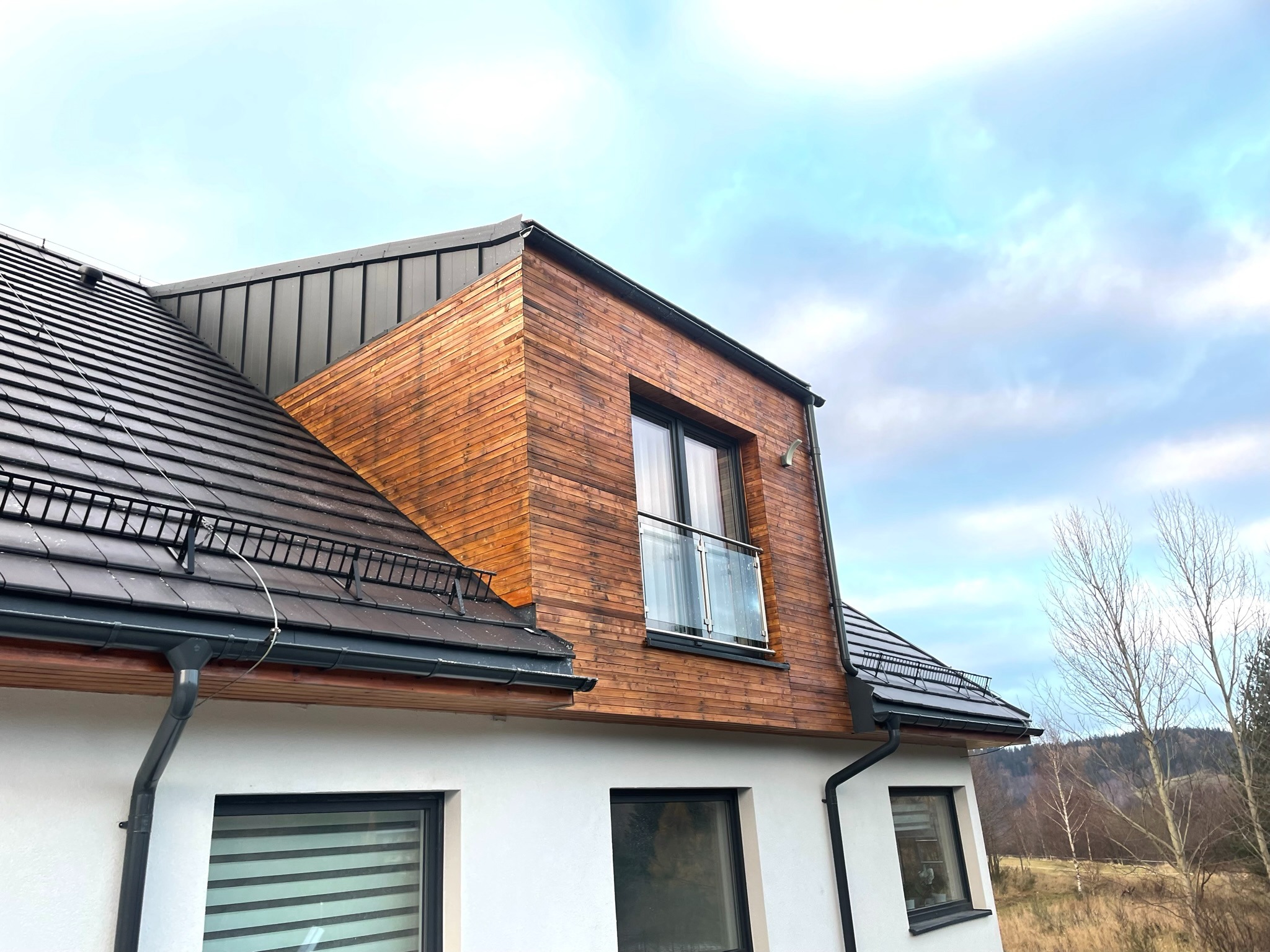 Nowoczesny dom z dachem pokrytym ciemną dachówką, drewnianą elewacją i balkonem ze szklaną balustradą, widok z dołu na tle pochmurnego nieba.