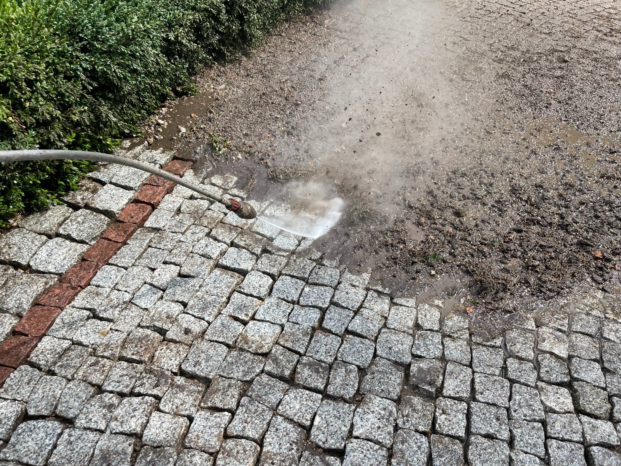 Dysza lancy spryskuje strumieniem wody pod ciśnieniem fragment szarej kostki brukowej, obok widoczny pas kostki w kolorze czerwonym, a dalej ziemia z resztkami roślinności.