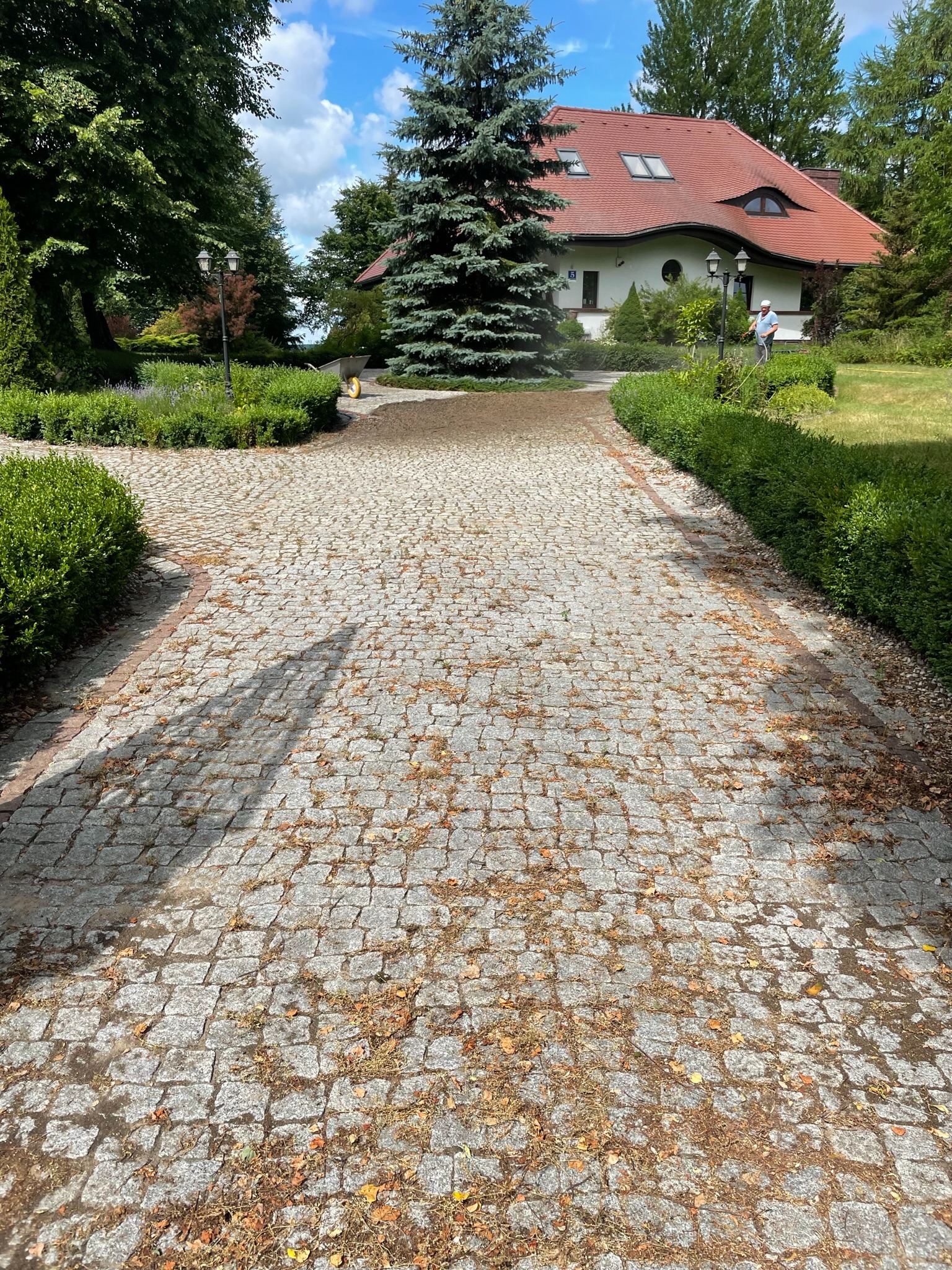Czyszczenie kostki brukowej z liści i zabrudzeń na posesji z domem jednorodzinnym; osoba w oddali używa węża ogrodowego.