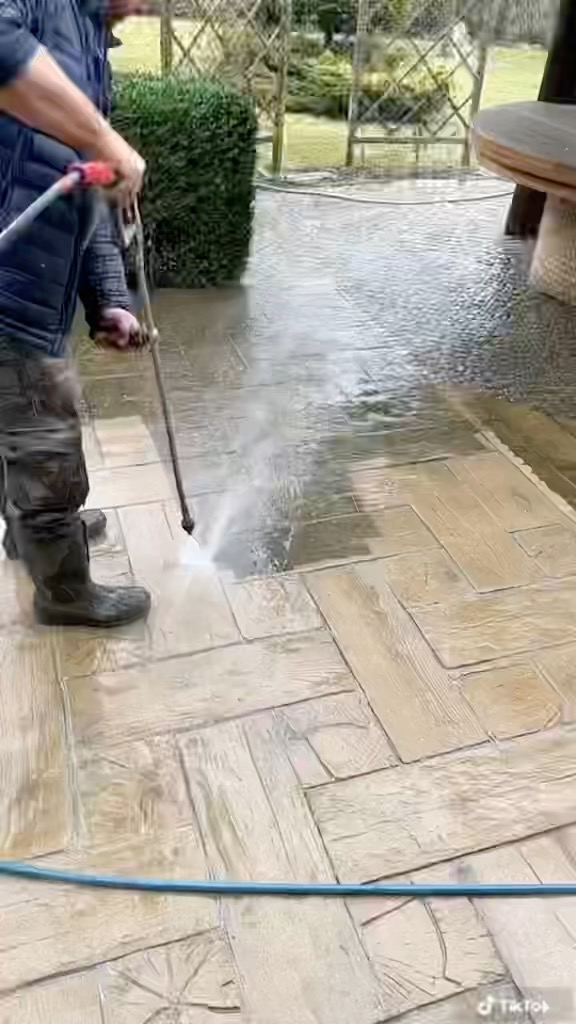 Czyszczenie kostki brukowej na zewnątrz domu za pomocą myjki ciśnieniowej; widoczna osoba w gumowych butach i kurtce, kierująca strumień wody na powierzchnię ułożoną w wzór jodełki.