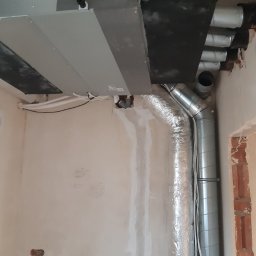 ColdWind Pawel Oszajca - Szare urządzenie centralne z podłączonymi rurami wentylacyjnymi w surowym wnętrzu, widoczne niedoskonałości ścian i brak wykończenia.