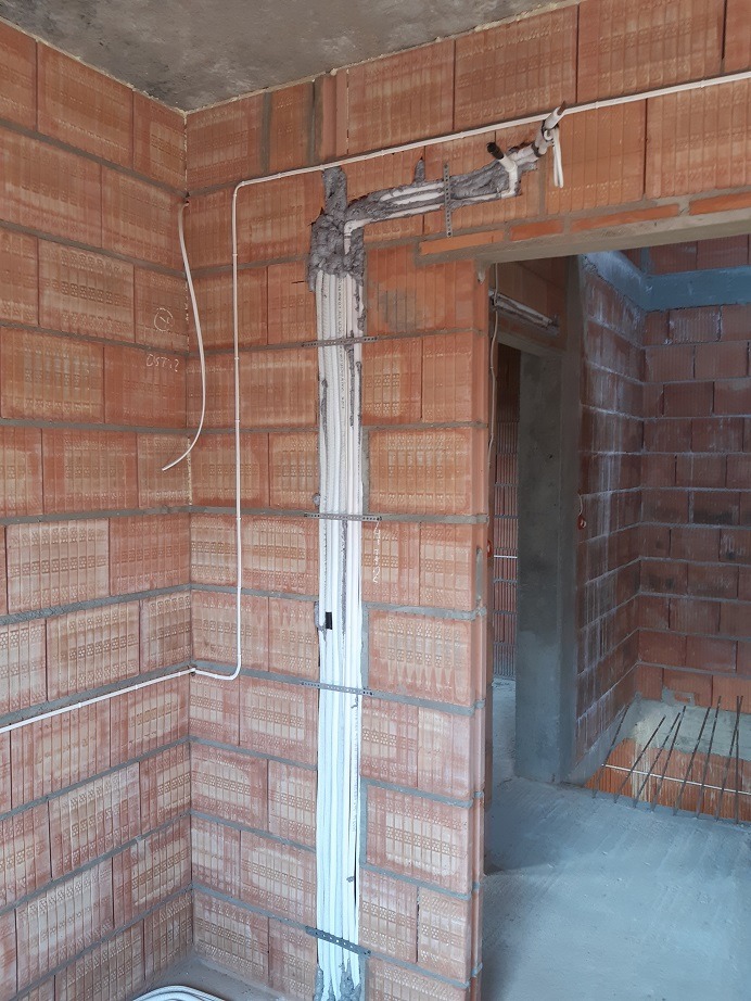 Instalacja przewodów wentylacyjnych w budynku w trakcie budowy, rury prowadzone pionowo po ceglanej ścianie, zamocowane metalowymi uchwytami, zabezpieczone pianką montażową w górnej części.