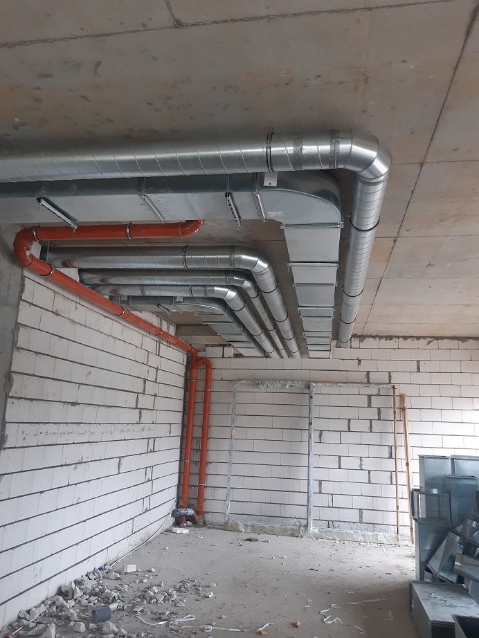 Surowe wnętrze w trakcie instalacji systemu przewodów wentylacyjnych i rekuperacyjnych, widoczne metalowe rury pod sufitem i pomarańczowe rury na ścianie z białego bloczka.