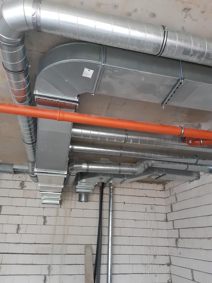 System kanałów wentylacyjnych z ocynkowanej blachy, zamontowany pod sufitem w surowym stanie, widoczne pomarańczowe rury i ściany z białego pustaka.