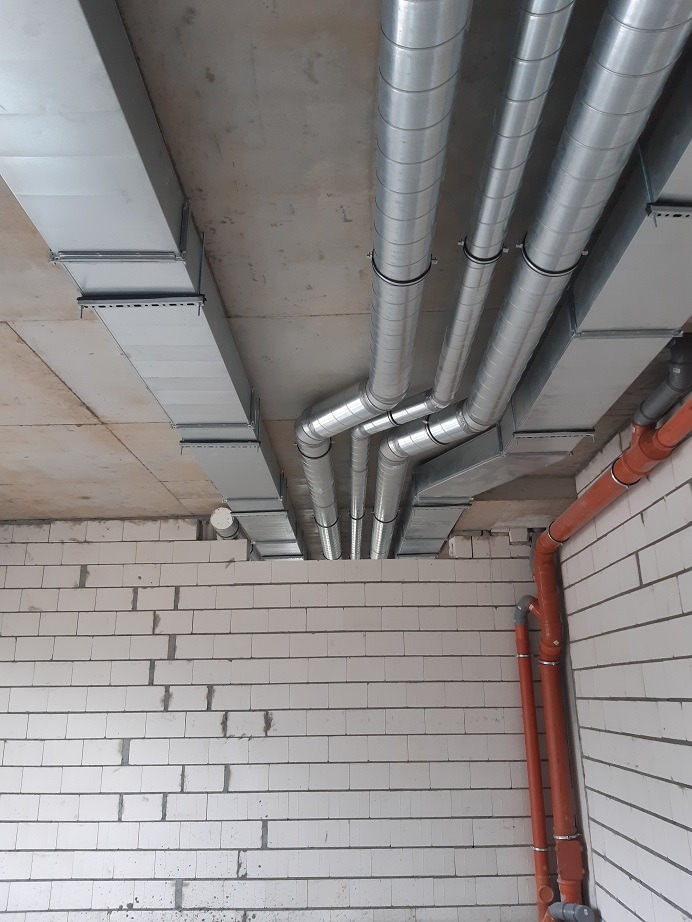 Instalacja przewodów wentylacyjnych: widok na sufit z widocznymi prostokątnymi i okrągłymi, metalowymi kanałami wentylacyjnymi, oraz pionowymi, pomarańczowymi rurami PCV na tle surowych, białych...