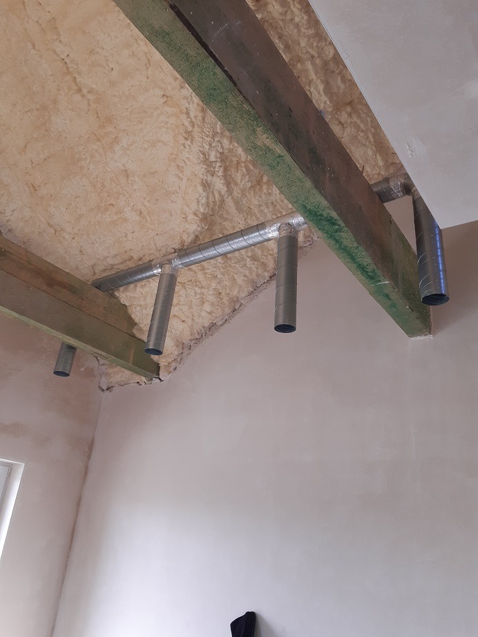 Montaż systemu wentylacji: widok na sufit z izolacją piankową i metalowymi rurami wentylacyjnymi przymocowanymi do drewnianych belek.