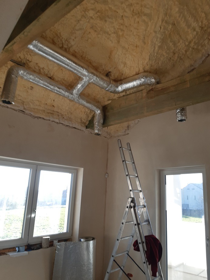 Montaż izolowanych kanałów wentylacyjnych w nowym budynku, widoczna drabina i materiały izolacyjne przy oknie.