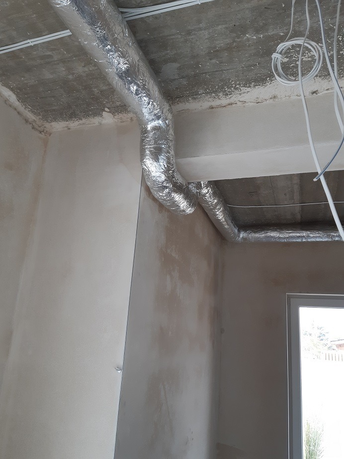 Rury izolowane folią aluminiową, prawdopodobnie system rozprowadzania powietrza, zamontowane pod surowym betonowym sufitem w pomieszczeniu w trakcie wykańczania, widoczne przewody elektryczne...