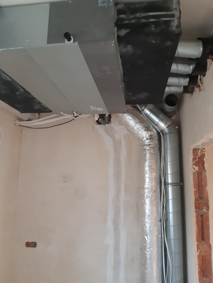 Szare urządzenie centralne z podłączonymi rurami wentylacyjnymi w surowym wnętrzu, widoczne niedoskonałości ścian i brak wykończenia.