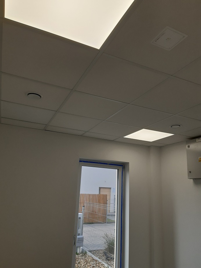 Białe, kasetonowe panele sufitowe z wbudowanym oświetleniem LED w pomieszczeniu biurowym, widoczny fragment ściany z oknem i instalacją elektryczną.