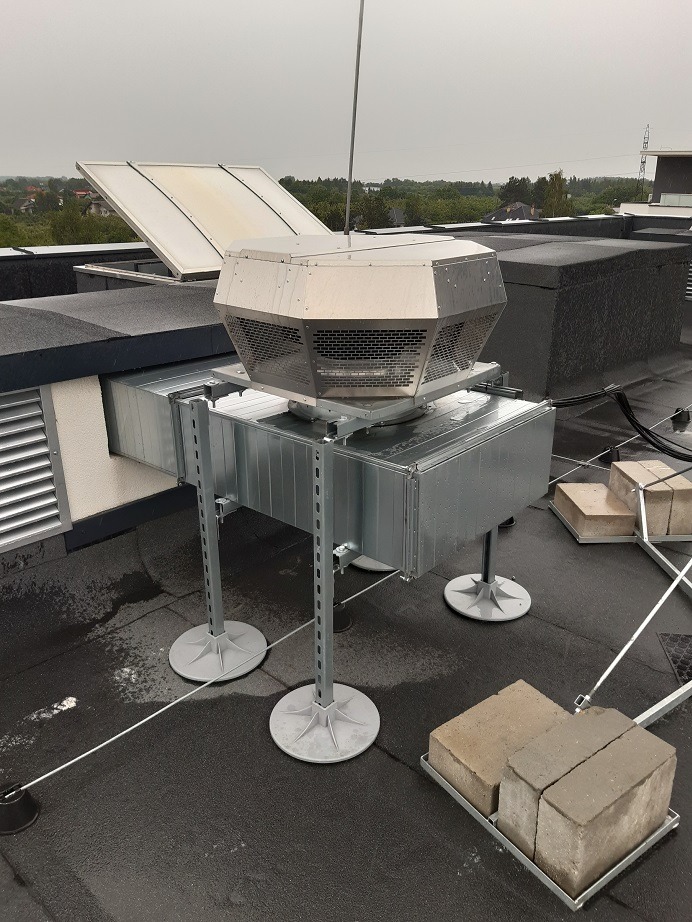 Metalowy system wentylacyjny z kratką ochronną, zamontowany na płaskim dachu, podparty na regulowanych wspornikach z obciążeniem z cegieł, widok z góry w pochmurny dzień.
