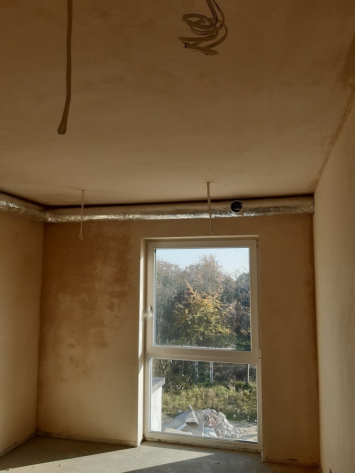 Wnętrze w stanie deweloperskim z widoczną instalacją wentylacyjną pod sufitem, rurami wystającymi z sufitu i oknem z widokiem na drzewa.