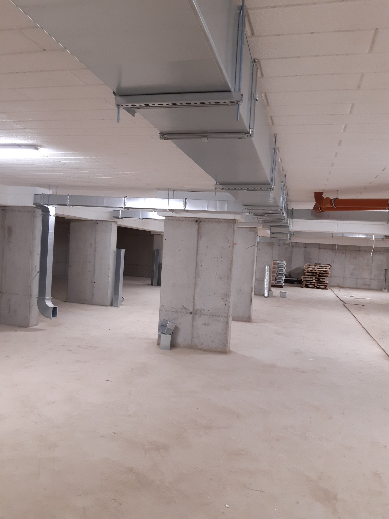 Szare, prostokątne kanały wentylacyjne zamontowane pod sufitem w surowym, betonowym pomieszczeniu. Pionowe rury wentylacyjne przy betonowych kolumnach. Widoczne palety w tle.