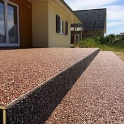 Czerwono-brązowa, kamienista nawierzchnia tarasu z dwoma stopniami, wykończona metalową listwą, przylegająca do budynku z elewacją w kolorze jasnej ochry i drewnianą ścianą, w tle widoczny dom...