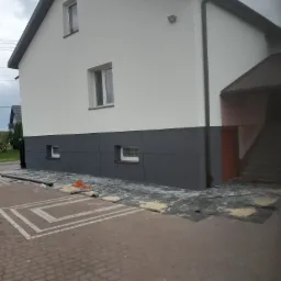 Dwukolorowa elewacja domu jednorodzinnego, dolna część w odcieniach szarości, górna w bieli, zabezpieczona folią podczas prac wykończeniowych.