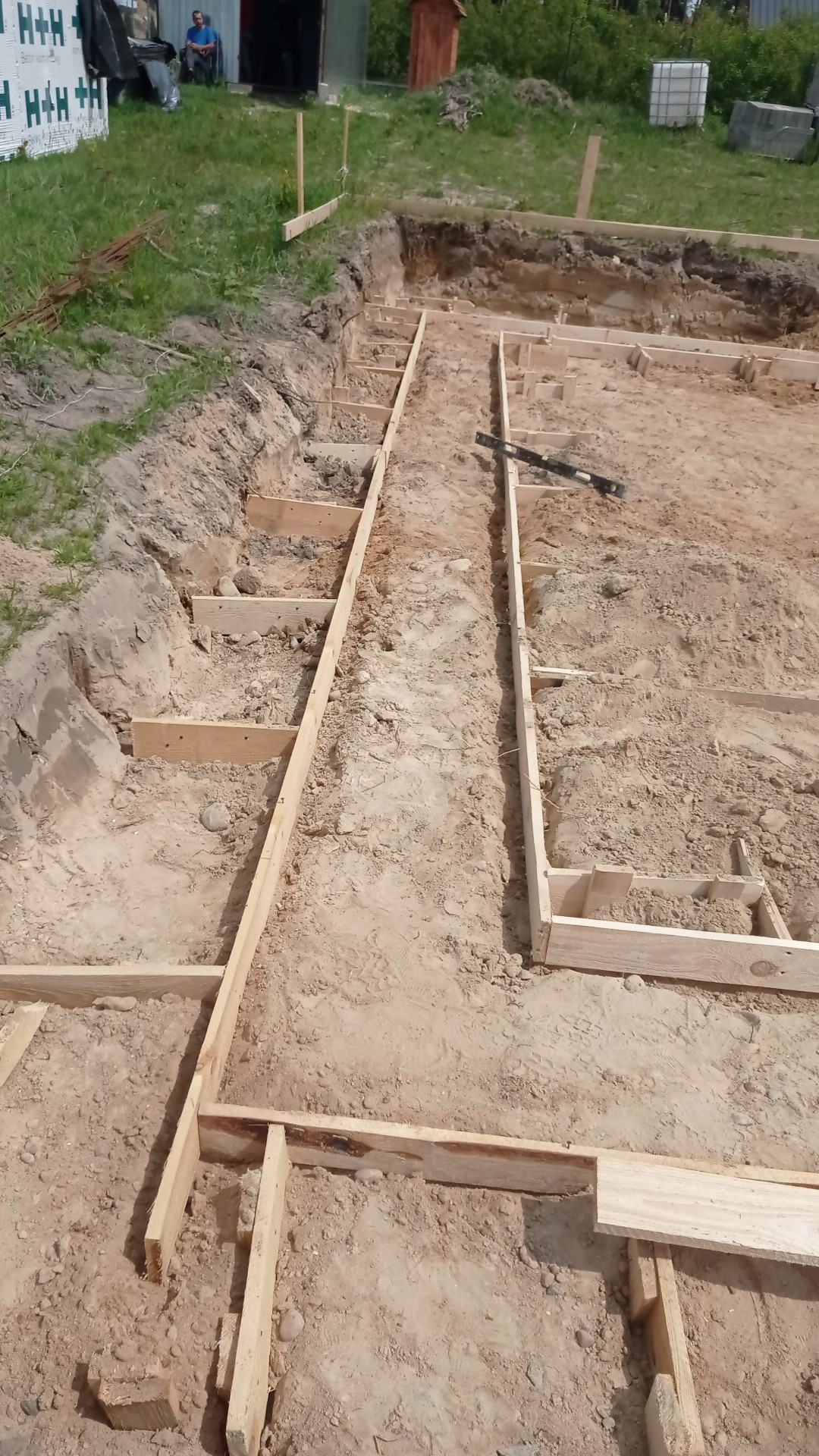 Przygotowanie drewnianego szalunku pod wylewkę fundamentów domu jednorodzinnego, widoczny poziomica na szalunku, w tle składowane bloczki H+H oraz budynek gospodarczy.
