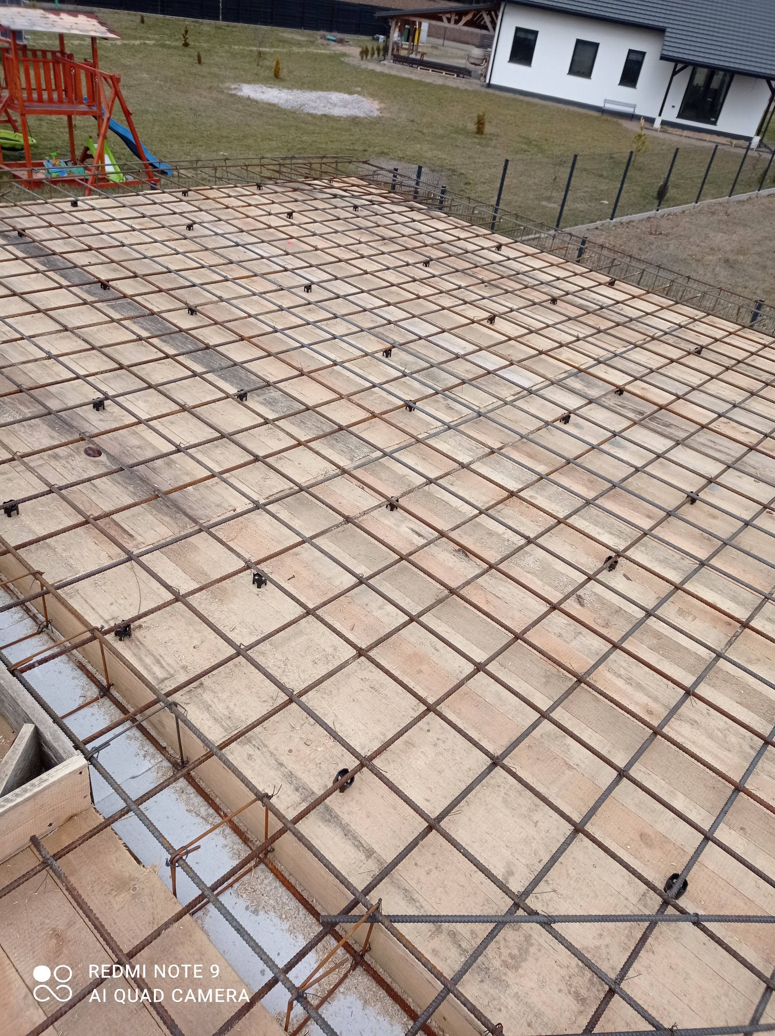 Siatka zbrojeniowa ułożona na deskowaniu szalunkowym, przygotowana do wylania fundamentu domu jednorodzinnego. W tle widoczny plac zabaw i budynek mieszkalny.