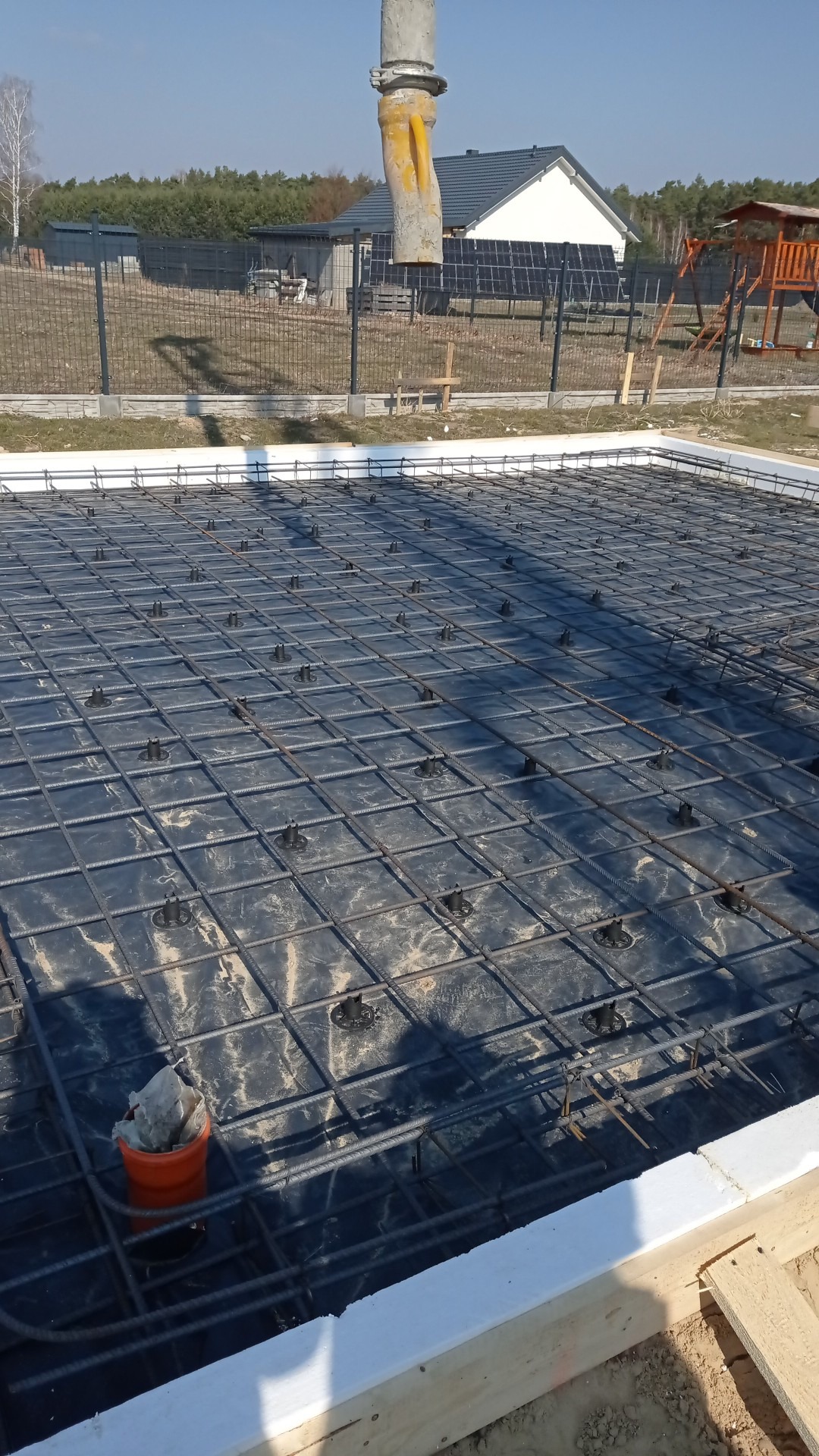 Przygotowanie zbrojenia płyty fundamentowej z folią i dystansami, widoczna pompa do betonu, w tle dom z panelami słonecznymi.