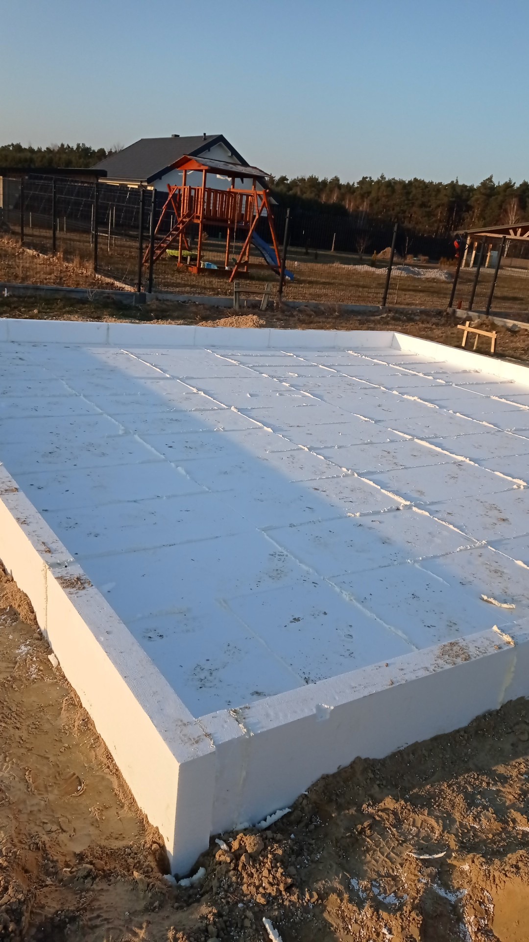 Fundament domu w trakcie budowy: białe bloczki izolacyjne ułożone na ziemi, przygotowane pod wylewkę, w tle plac zabaw i dom jednorodzinny.
