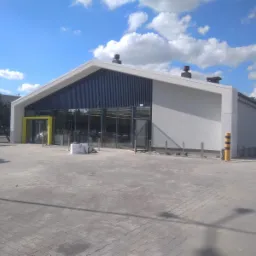 Nowoczesny budynek handlowy z jasnoszarą elewacją, ciemnoszarymi pionowymi panelami i żółtymi ramami drzwi wejściowych, widoczny z zewnątrz, na placu budowy z materiałami budowlanymi i sprzętem.