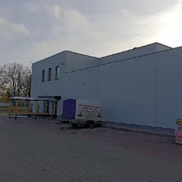 Świeżo pomalowana, jasnoszara elewacja budynku z trzema oknami, widoczny przyczepka z reklamą wypożyczalni sprzętu budowlanego i paleta z workami materiałów budowlanych.