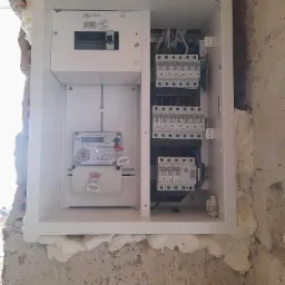 Wnętrze skrzynki elektrycznej z zamontowanym licznikiem jednofazowym, bezpiecznikami i przewodami, osadzone w surowej ścianie z widoczną pianką montażową.