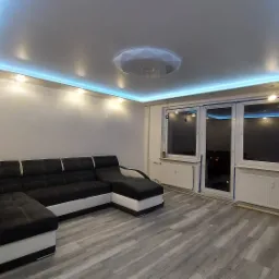 Salon po remoncie z szarymi panelami podłogowymi, narożnikiem w odcieniach szarości i bieli oraz sufitem podwieszanym z niebieskim oświetleniem LED.