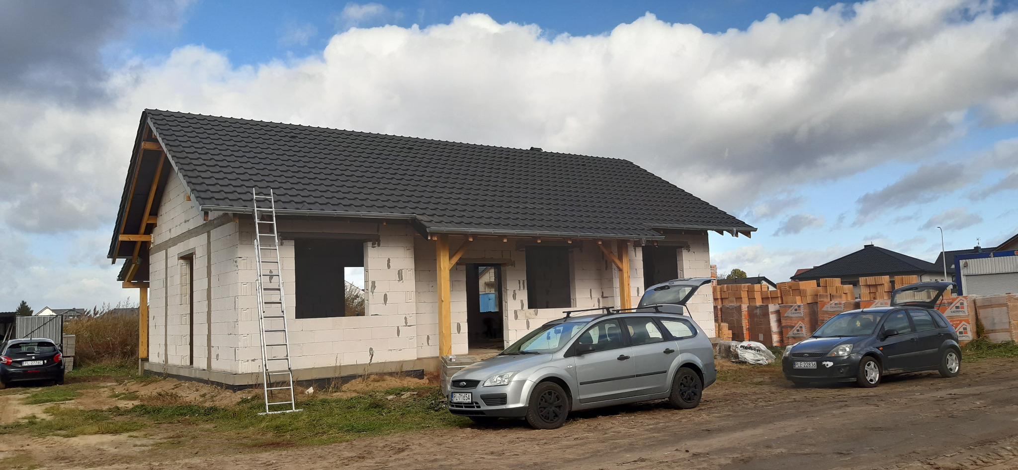 Dom w trakcie budowy z nowym, ciemnym dachem z dachówki ceramicznej, widoczne rusztowanie i przygotowane okna, na podwórku zaparkowane samochody.