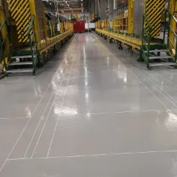 Jasnoszara, błyszcząca posadzka w hali produkcyjnej, z białymi liniami wyznaczającymi pola, widoczne żółto-czarne platformy robocze z zielonymi schodkami po bokach.