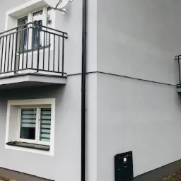 Szary dom z dwoma balkonami z czarnymi, metalowymi balustradami, widok z zewnątrz.